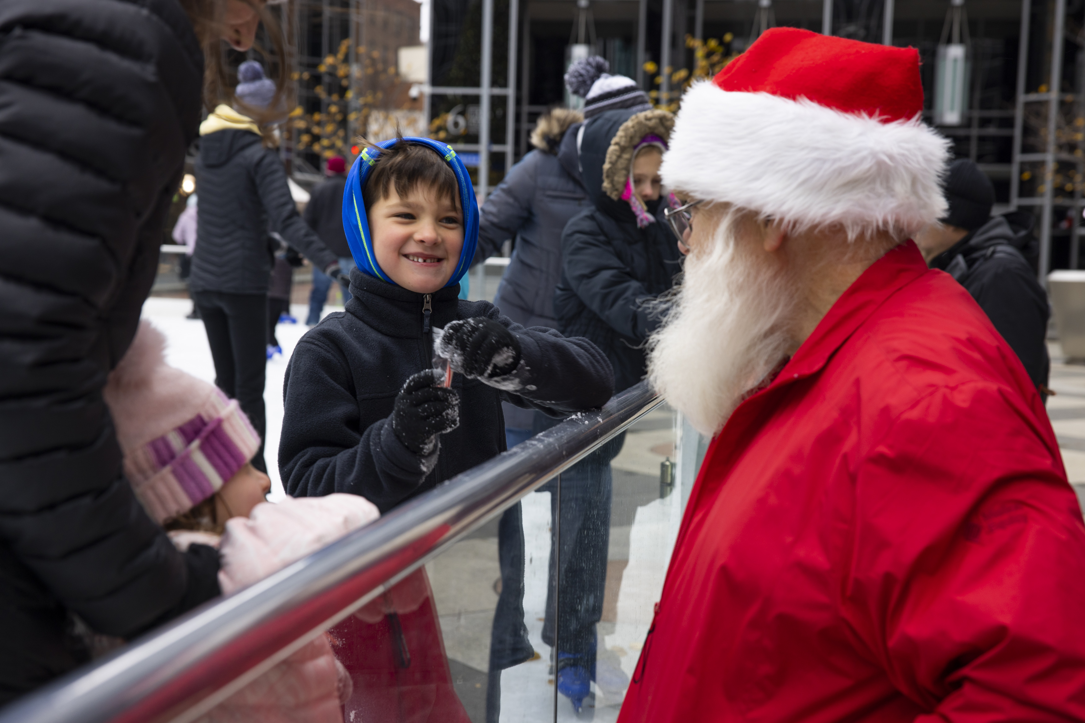 Ken Barker, an older, white man is dressed in a red Santa suit, talks to children at an outdoor ice skating rink. 